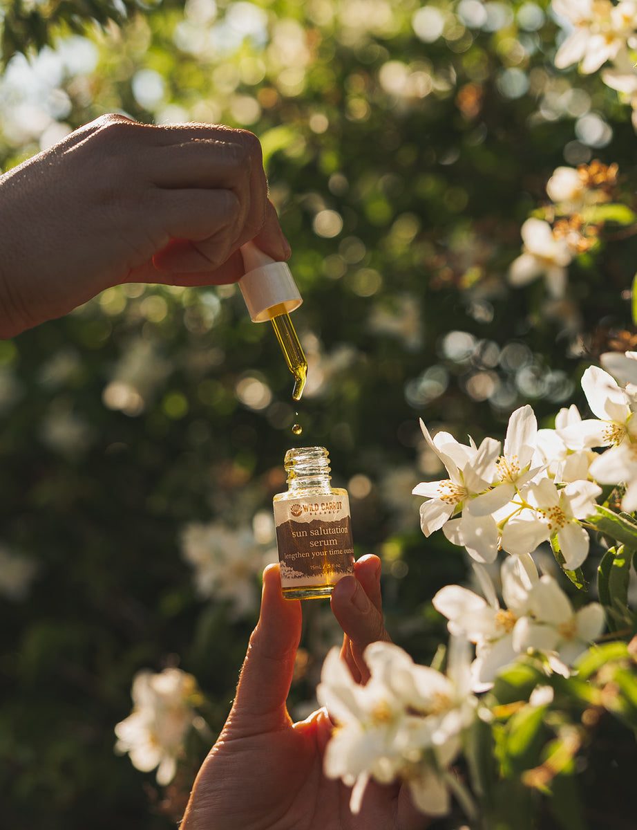 Hand holding a dropper with a small bottle of oil, surrounded by white flowers and greenery.